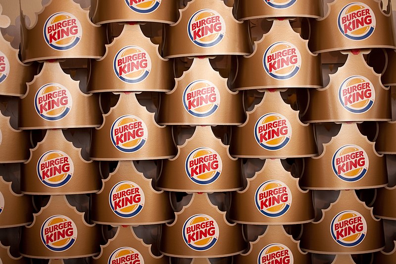 Stacks of cardboard crowns featuring the Burger King logo are neatly arranged. The crowns are golden brown with the red and blue Burger King logo in the center, creating a repetitive pattern throughout the image.