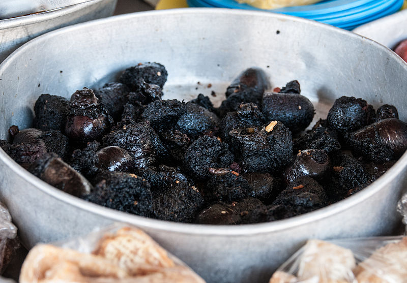 A large silver bowl filled with dark, charred-looking food pieces resembling cooked blood sausages or morcilla. The pieces have an uneven texture and appear crispy on the outside. There are other food items, wrapped in plastic bags, visible around the edge of the bowl.