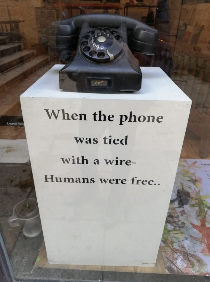 A vintage black rotary phone is displayed on a white pedestal inside a glass case. The pedestal has the text: "When the phone was tied with a wire - Humans were free.." The background shows a few items including papers on the floor and a partial staircase.