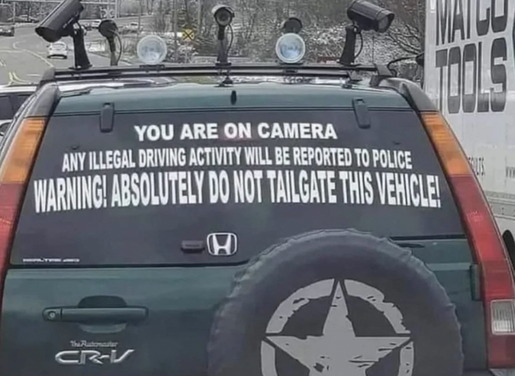 A green Honda CR-V with a prominent warning on the rear window stating: "YOU ARE ON CAMERA. ANY ILLEGAL DRIVING ACTIVITY WILL BE REPORTED TO POLICE. WARNING! ABSOLUTELY DO NOT TAILGATE THIS VEHICLE!" The car has roof-mounted lights and a spare tire on the back.