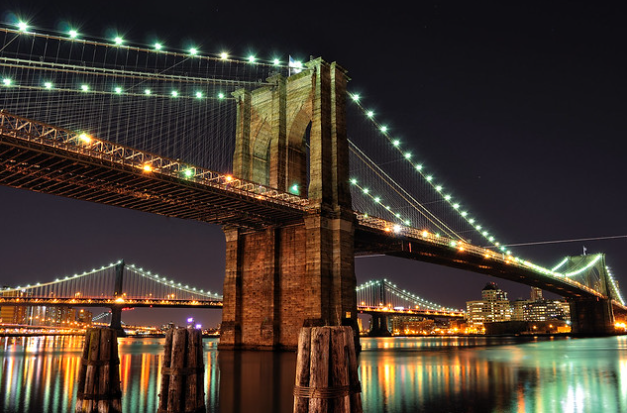 A nighttime view of the Brooklyn Bridge in New York City, illuminated with lights. The bridge spans across the water, with another lit bridge visible in the background. Reflective city lights and buildings are seen in the distance.