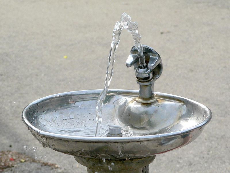 A close-up view of a stainless steel drinking fountain with water arching out of the spout. The fountain is outdoors, and the background is a concrete surface. Droplets of water are visible on the basin.