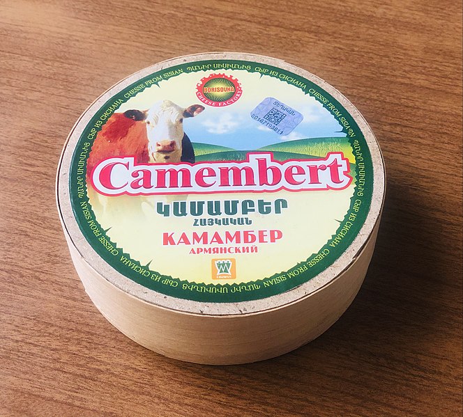 A round wooden box of Camembert cheese with a green-rimmed lid featuring a cow image and text in Armenian and Russian. The label has a mountain and field background and depicts classic Camembert packaging design. The box is placed on a wooden surface.