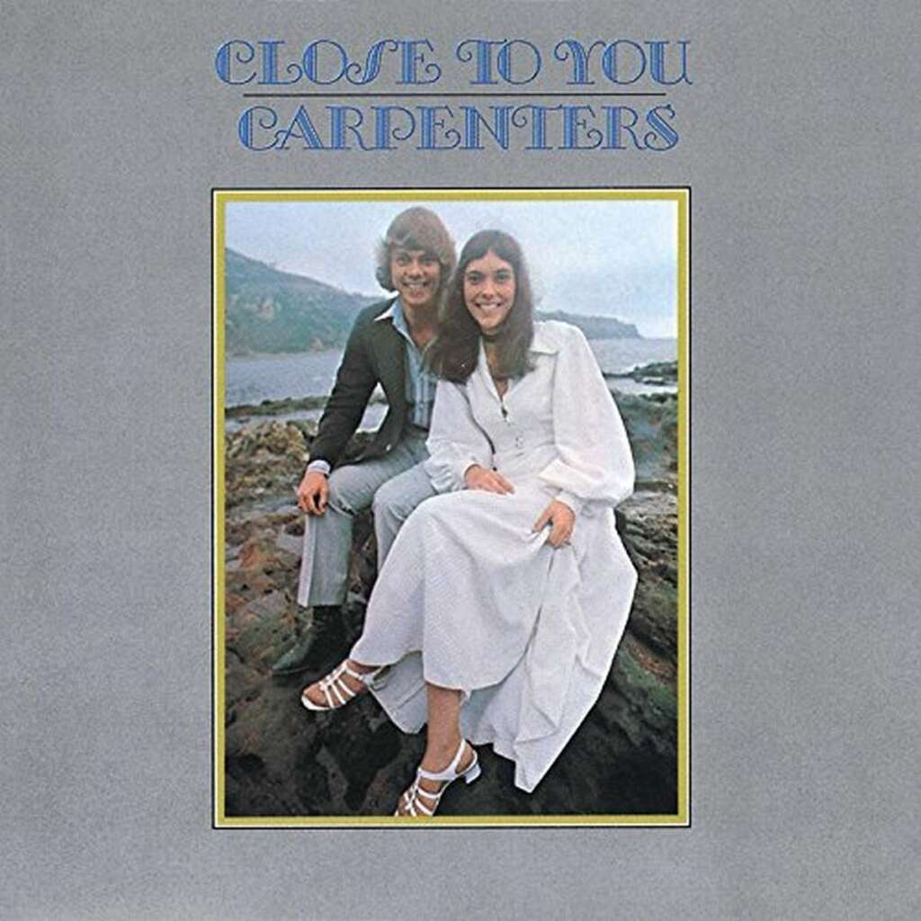 An album cover titled "Close to You" by Carpenters features a man with blond hair in a gray suit and a woman with long dark hair in a white dress sitting on rocky terrain by the sea, smiling. The title is written in blue uppercase letters at the top.