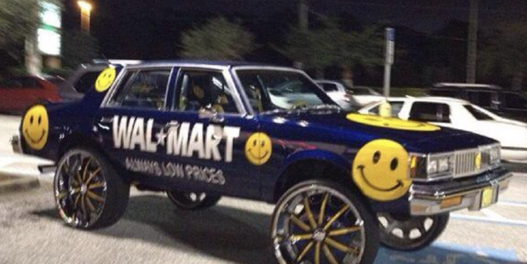 A modified car in a parking lot features oversized wheels, a blue body, and Walmart branding, including large smiley face decals and the slogan "Always Low Prices." The car is outfitted with multiple Walmart logos, and it is nighttime.