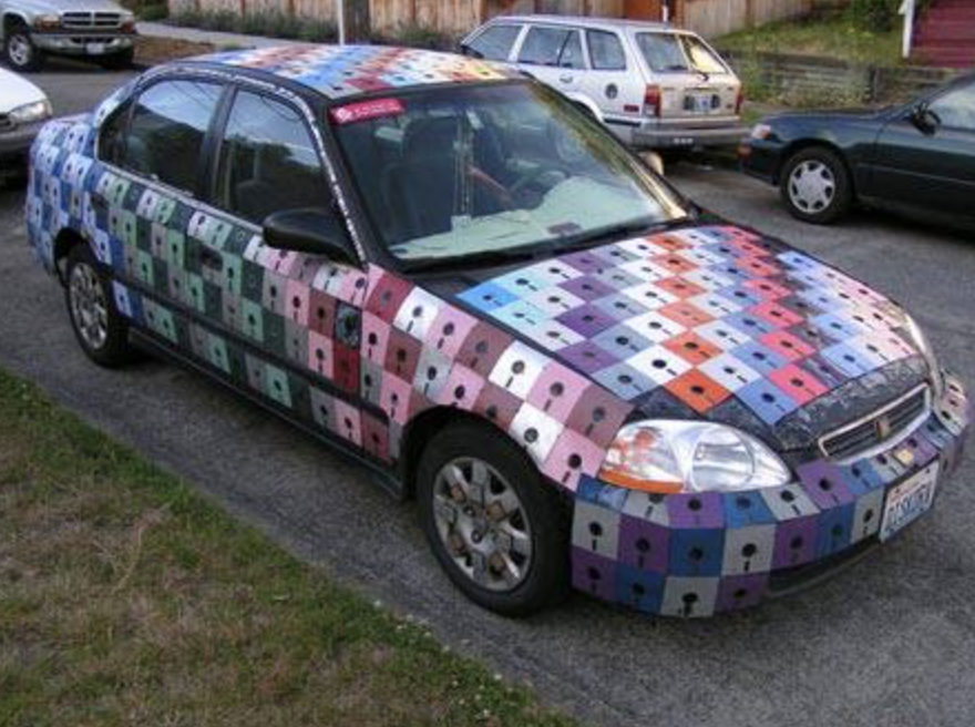 A car is covered in a colorful mosaic pattern made up of small squares resembling floppy disks. The design covers the entire exterior, including the hood, roof, and doors. The car is parked on a residential street alongside other vehicles.