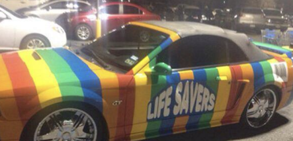 A colorful car is painted with rainbow stripes and the word "LIFE SAVERS" emblazoned on the door, mimicking the famous candy packaging. The car is parked in a lot at night, with other vehicles visible in the background.