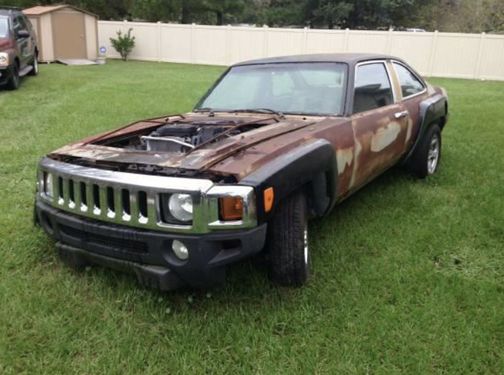 An old car with a rusted body is parked on a grassy lawn. The front of the car appears to be modified with the grille and bumper of a Hummer, while the rest of the body is a different, older model. The hood is missing, exposing the engine compartment. A fence and a shed are in the background.