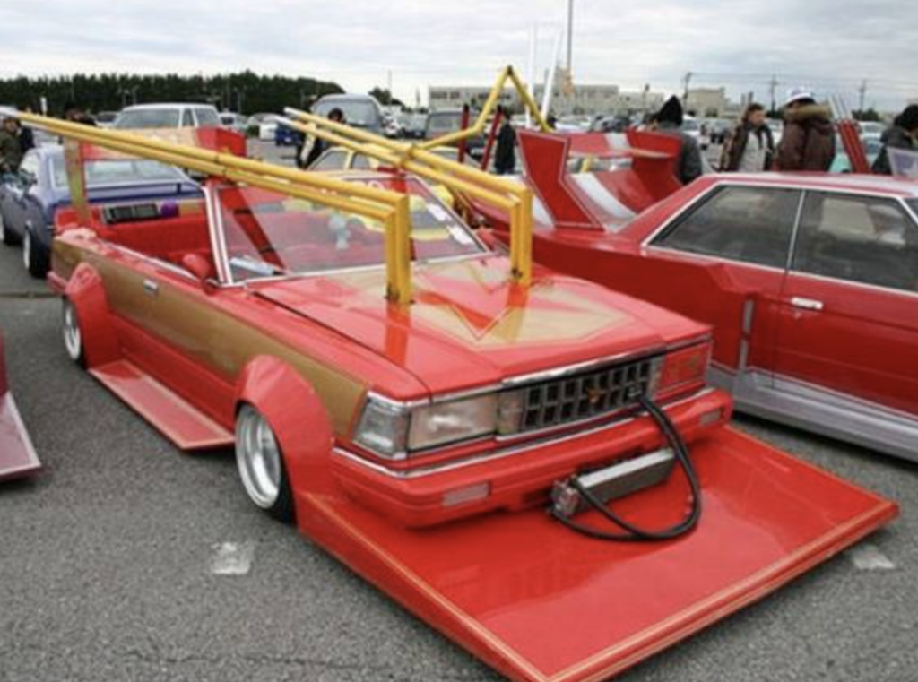 A modified red car with a low ground clearance, wide body kit, and exaggerated features like a large front splitter and yellow roll bars. The car is parked among other custom vehicles in an outdoor lot, with people in the background.