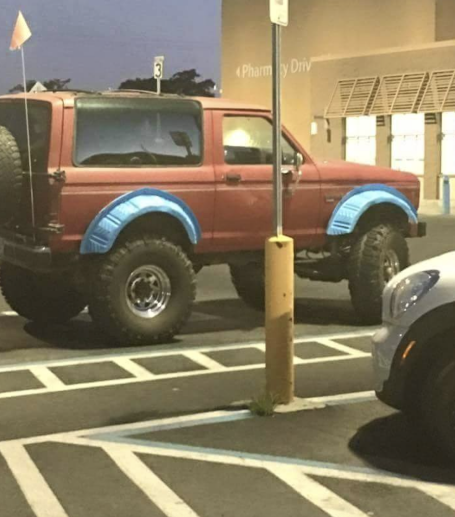 A red SUV with oversized tires is parked in a handicapped parking space without a permit. Blue tape is visible around the wheel arches. A silver car is partially visible in the bottom right corner of the image.