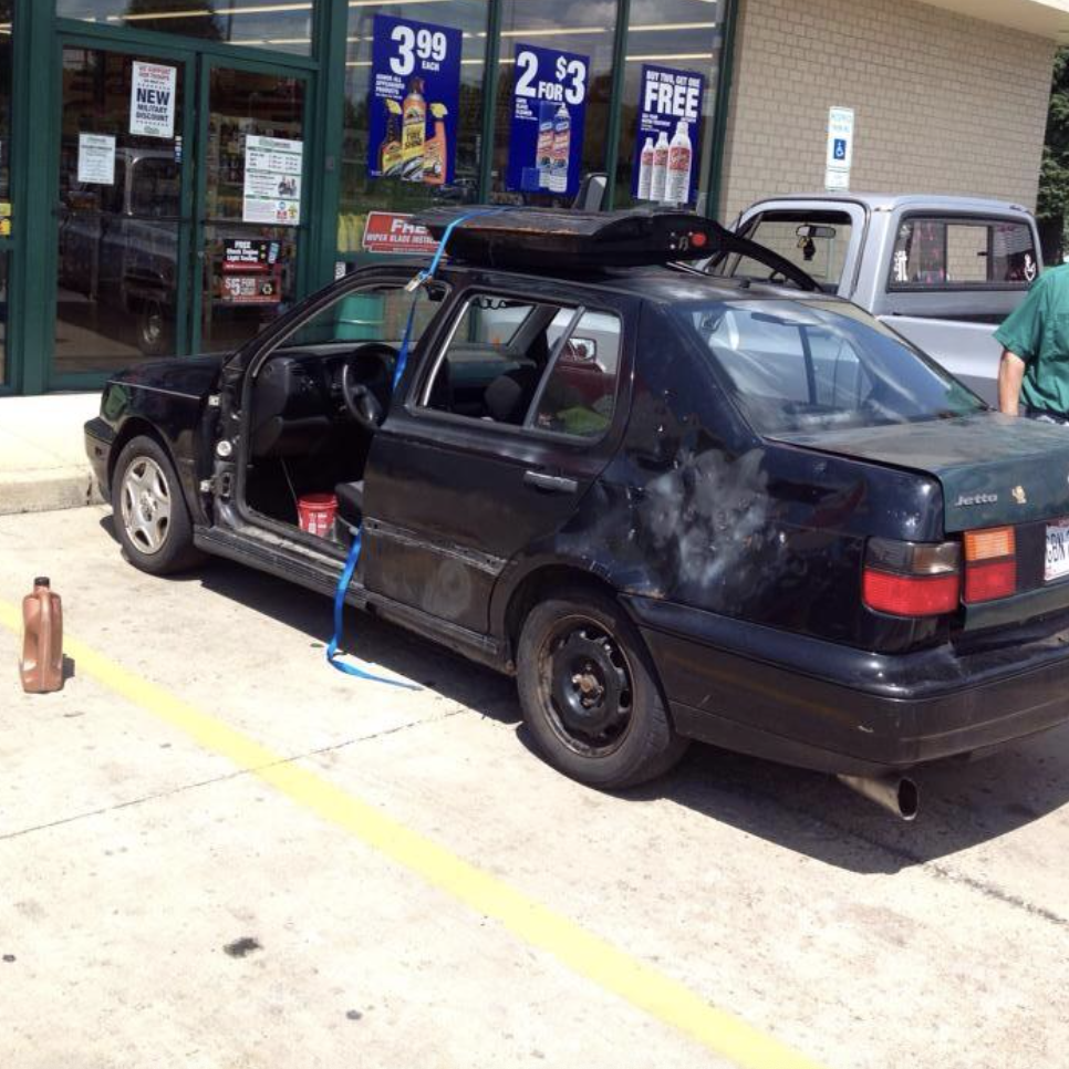 A black sedan with its driver's side door open is parked in front of a convenience store. The car appears to be undergoing maintenance, with a canister of motor oil and another container placed nearby on the ground. A person in a green shirt is partially visible.