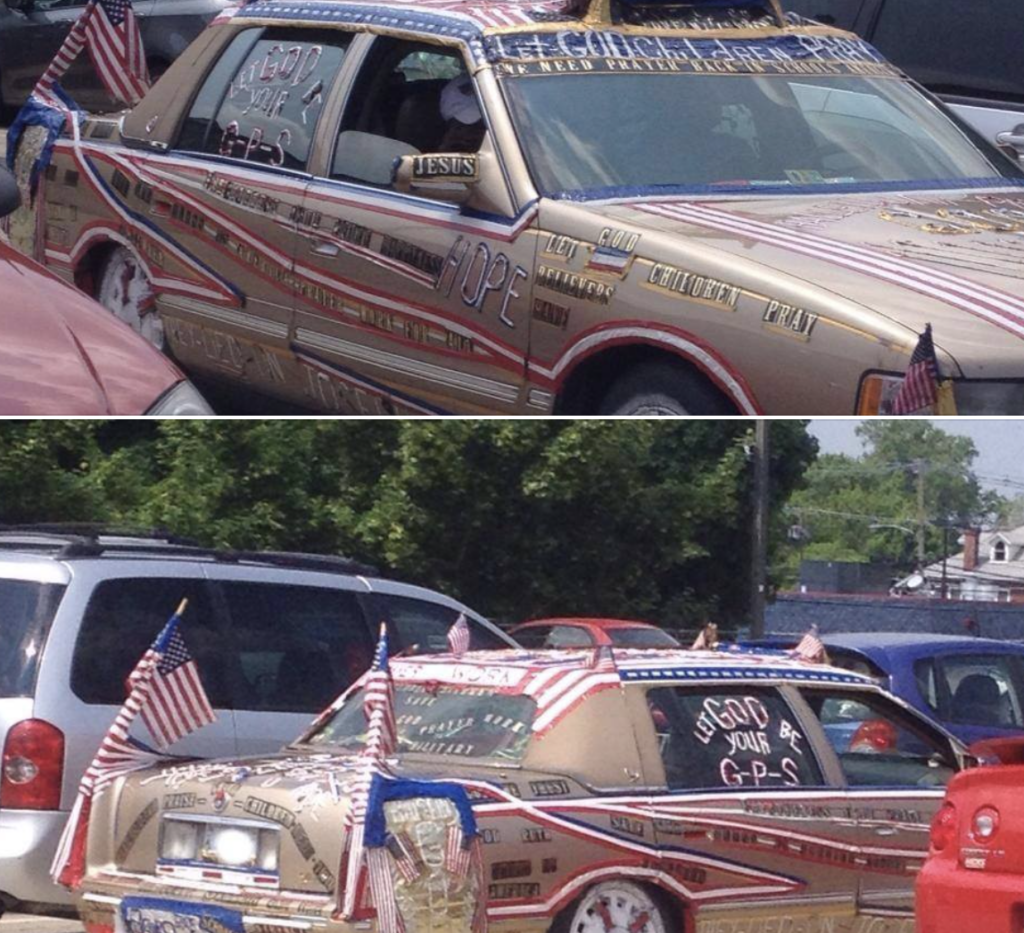 A tan sedan intricately decorated with numerous messages, flags, and religious symbols is parked in a lot. The car is covered in text proclaiming themes of hope, prayer, and Jesus, with American flags mounted on different parts of the vehicle.