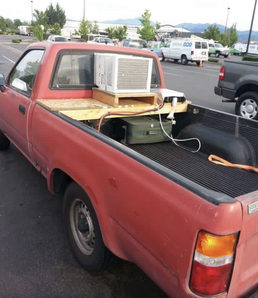 A red pickup truck is parked in a lot. An air conditioning unit is installed in the back on a wooden platform, plugged into what appears to be a portable generator. Other cars and a building are visible in the background.