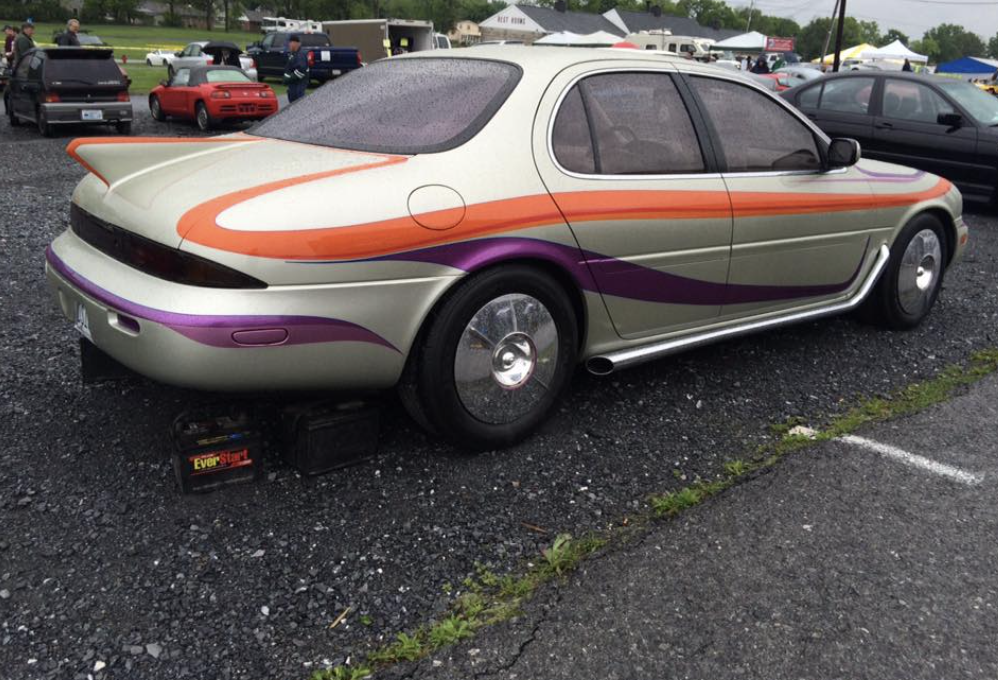 A customized car with a streamlined design featuring a beige body with orange and purple stripes and accented with large chrome hubcaps. The car is parked on gravel, with miscellaneous items and vehicles visible in the background.