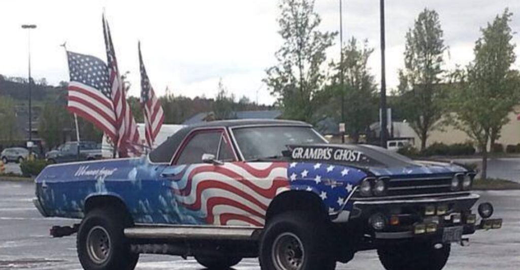 A lifted classic car with large tires, decorated with a patriotic red, white, and blue American flag theme, including three American flags mounted at the rear. The car's hood displays the phrase "Gramps Ghost." The car is parked in a nearly empty lot.