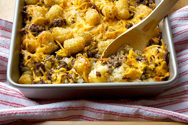 A close-up of a baking pan filled with a tater tot casserole, featuring tater tots, ground beef, melted cheese, and various seasonings. A wooden spoon is scooping out a portion. The pan rests on a red and white striped cloth.