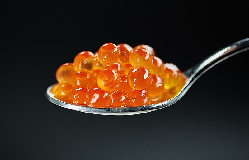 A close-up shot of several orange fish roe, known as salmon caviar, piled on a silver spoon. The transparent roe has a shiny appearance and is set against a solid black background, highlighting their vibrant color and glossy texture.