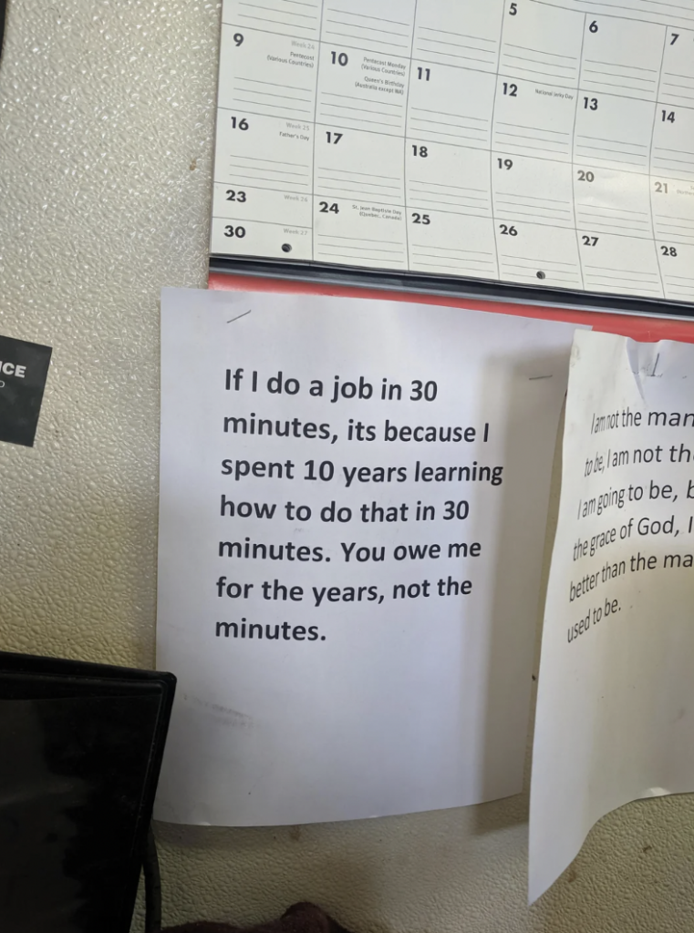 A wall displays two printed notes under a calendar. One note reads: "If I do a job in 30 minutes, it's because I spent 10 years learning how to do that in 30 minutes. You owe me for the years, not the minutes." The calendar shows blurred dates and numbers.
