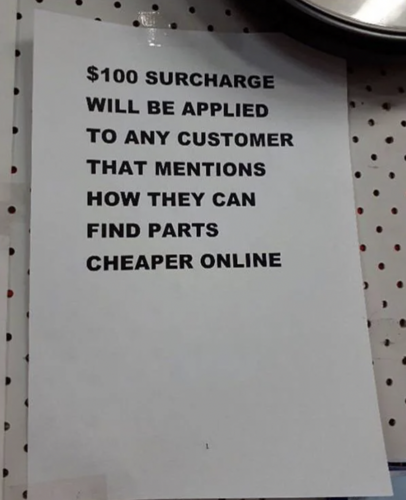 A white sign with bold black text reads: "$100 surcharge will be applied to any customer that mentions how they can find parts cheaper online." The sign is taped to a perforated board.