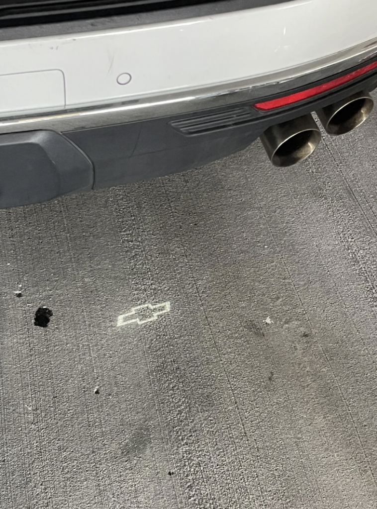 Close-up view of a car's rear bumper with dual exhaust pipes. A projection of the Chevrolet logo is visible on the ground beneath the car. The car is parked on a concrete surface.