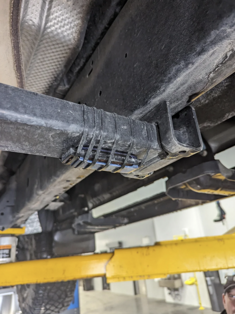 A close-up view of the underside of a vehicle, showing a black component attached to a metal bar. The vehicle is elevated on a yellow car lift. The background includes parts of the car lift and the workshop, which appears to be well-lit.