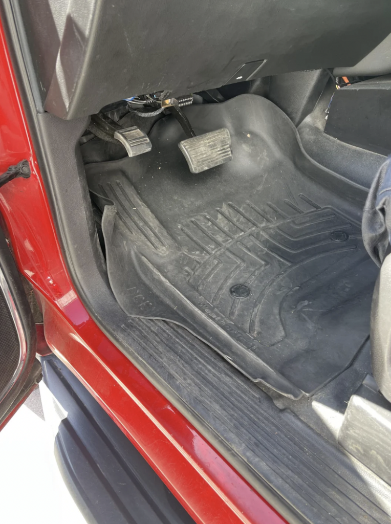 A close-up view of the driver's side footwell in a red vehicle, showing the black rubber floor mat, brake and accelerator pedals. The mat has grooves and dirt on it. The car door is open, and part of the car seat is visible on the right.