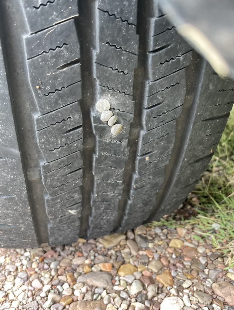 Close-up of a car tire tread with four partially embedded metal screws. The screws are arranged in a line, penetrating the rubber surface. Gravel and grass are visible on the ground beneath the tire.