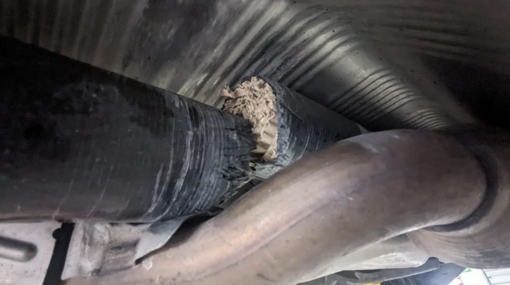 A close-up view of a damaged section of a car's exhaust pipe. The pipe appears to be split or broken, revealing a rough, irregular interior. The surrounding area includes other parts of the vehicle's undercarriage.