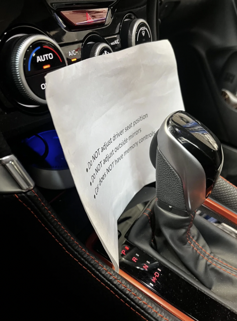A white paper sign is taped near the car's gear shift. The sign has printed text on it. Below the sign are the gear shift, wrapped in black leather with red stitching, and part of the car's center console, including the gear indicator.