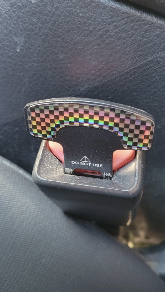Close-up of a car seatbelt buckle slot with a decorative, holographic checkered pattern insert. The insert has a label that reads "DO NOT USE," indicating it is not suitable for securing a seatbelt. The surrounding interior features black upholstery.