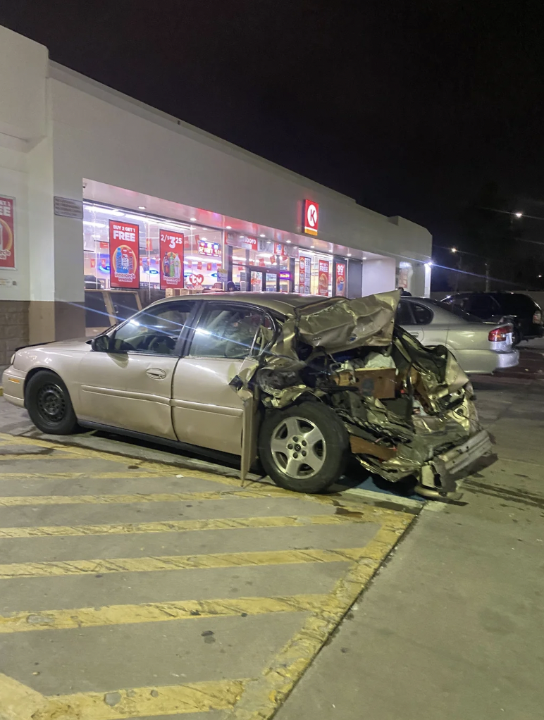 A gold sedan with significant rear-end damage is parked diagonally in a lot outside a convenience store at night. The car's trunk area is heavily crumpled, exposing internal components. The store has bright signage and displays offers in its windows.