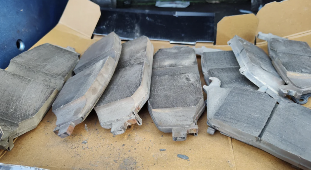 A set of six worn and dusty car brake pads are lying on a piece of cardboard. The pads show signs of significant wear with visible scratches and dirt, indicating they have been used extensively and may need replacement.