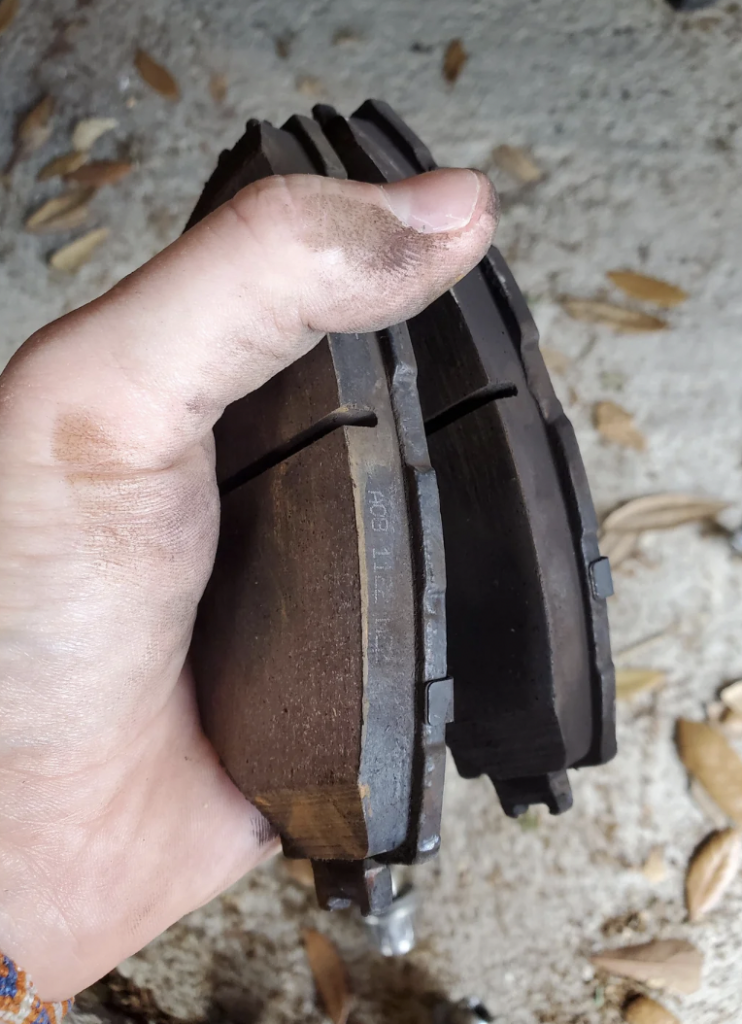 A hand holding two worn, dark-colored brake pads. The pads show visible wear and dirt, indicating they need replacement. The background is a rough, sandy surface with scattered dry leaves, and the person’s fingers appear slightly dirty.
