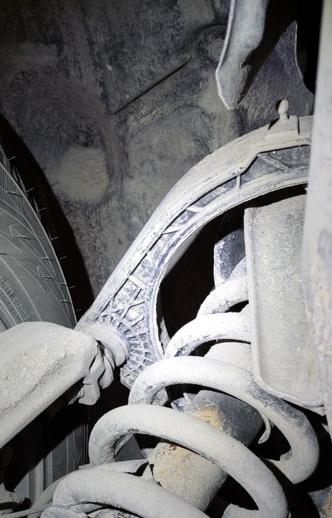 Close-up of a vehicle's undercarriage, showing a dusty and worn suspension component, including a shock absorber coil spring and part of a tire. The overall condition of the metal parts appears aged and corroded.