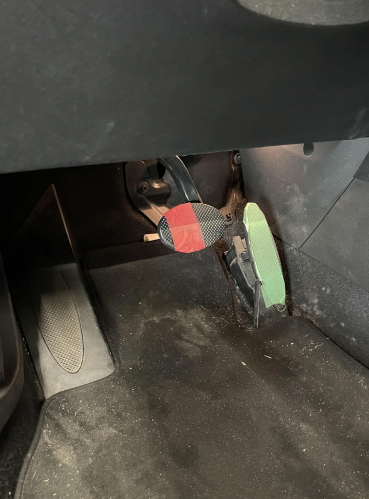 A close-up view of a car's foot pedal area. The brake pedal is taped with red tape and is located in the center, while the accelerator pedal, marked with green tape, is on the right. The floor appears slightly dirty with some dust and debris visible.