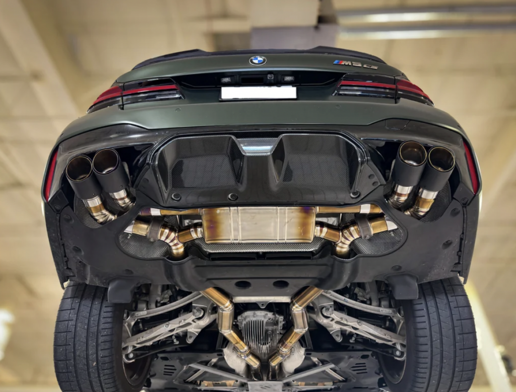 A view of the underside of a BMW M5 CS, focusing on the car's detailed exhaust system and mechanics. The car is elevated, showing quad exhaust tips, a large performance exhaust system, and part of the rear suspension components.