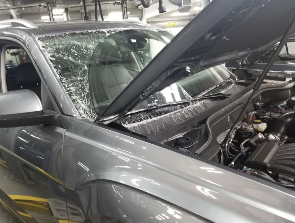 A silver SUV with significant windshield damage and the hood open, revealing the engine bay. The windshield is shattered, and glass pieces are spread on the car's hood. The vehicle is inside an automotive repair shop with visible tools and equipment in the background.