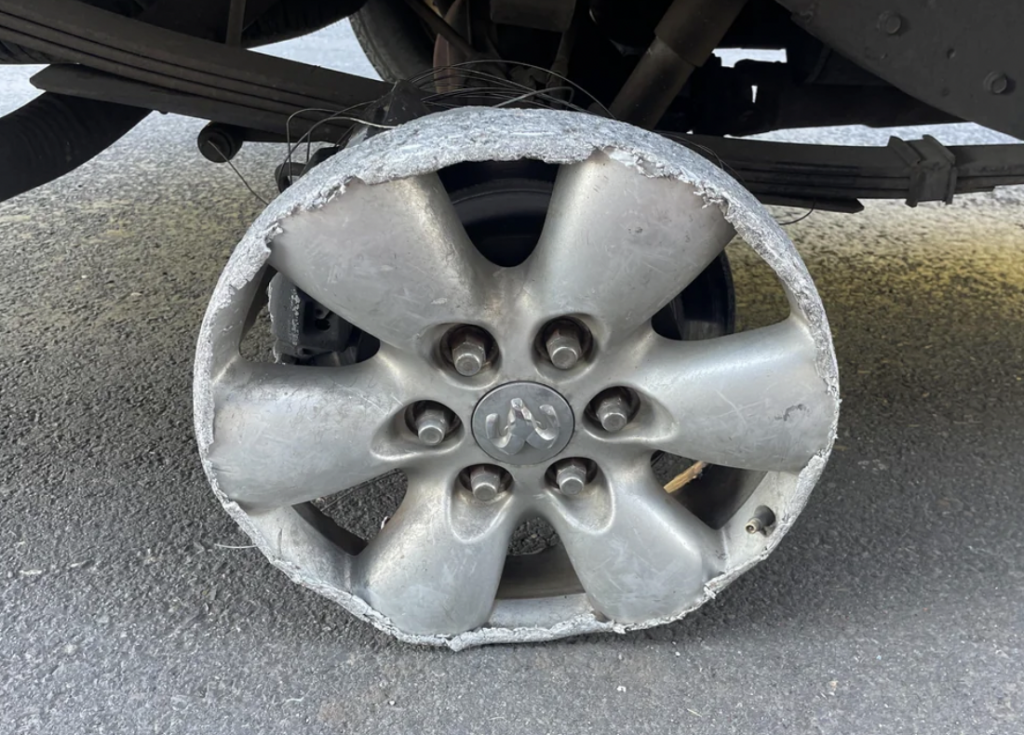Close-up view of a severely damaged car wheel, with the tire completely shredded away, exposing the metal rim, which appears worn and corroded. The wheel is partially suspended off the ground beneath a vehicle, highlighting the extent of the damage.