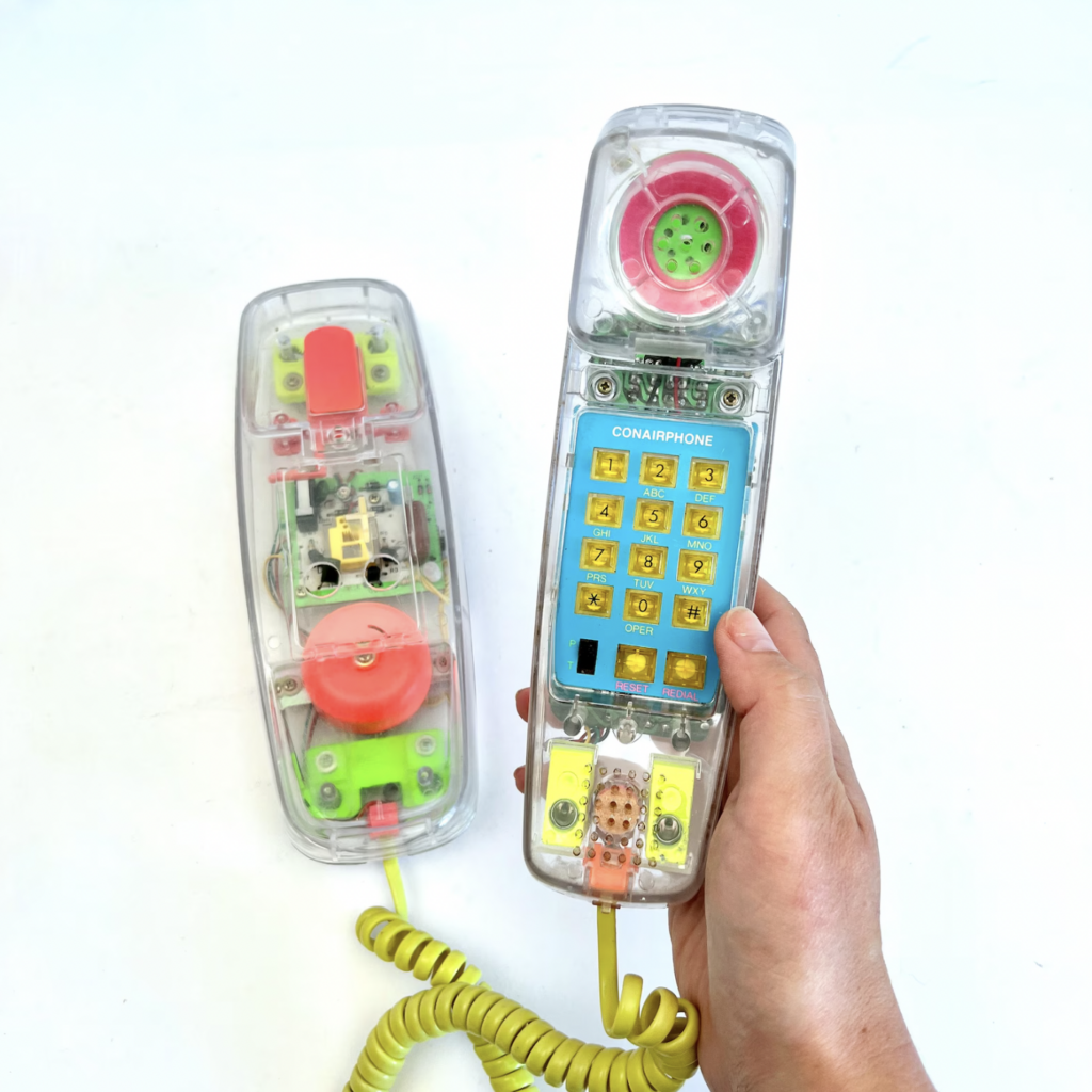A person holding a vintage clear plastic toy phone with visible internal components. The phone features colorful buttons and wiring, with a yellow coiled cord. The detached base of the phone lies next to it on a white background.