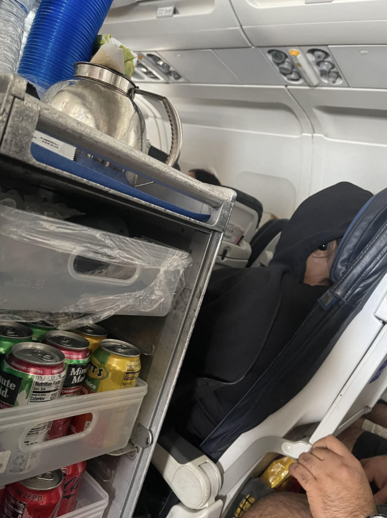 A beverage cart containing various canned drinks and a metal teapot is seen in the aisle of an airplane. An individual wearing a black hoodie and glasses is seated in a nearby seat, with other passengers visible in the background.