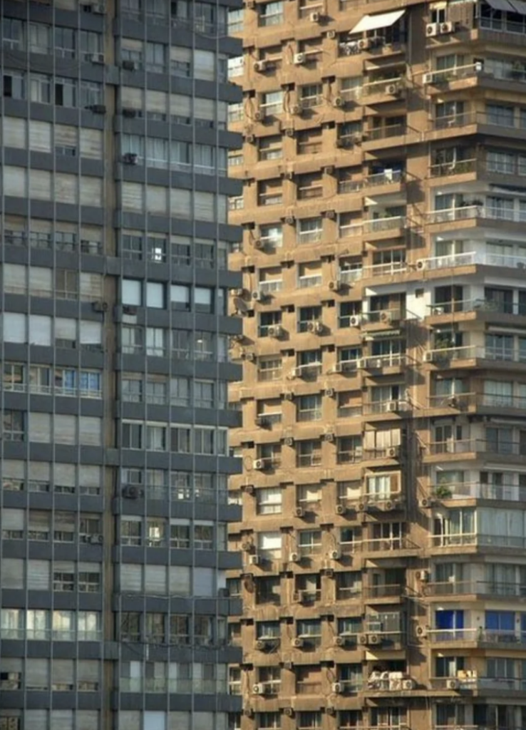 A close-up view of two tall, densely populated apartment buildings. The building on the left is grey with repetitive window patterns, while the one on the right is brown with numerous balconies, air conditioning units, and various items visible on the balconies.