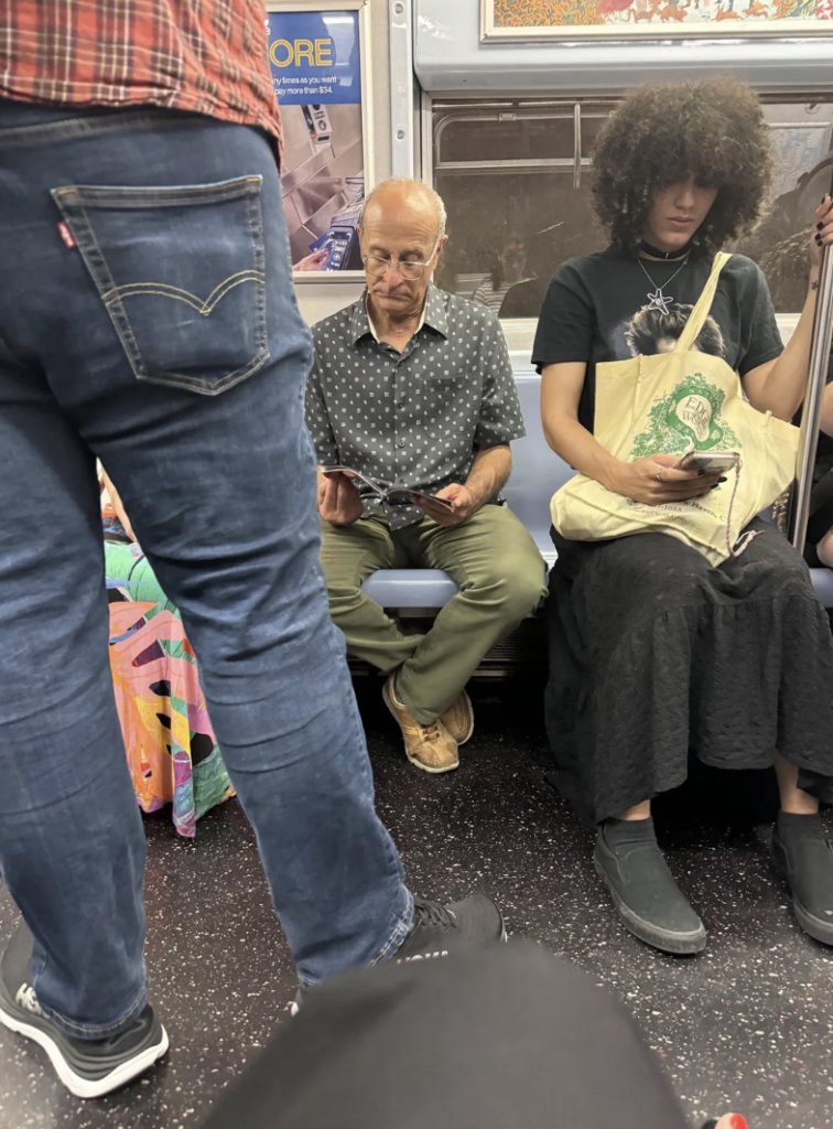 A passenger wearing a dotted shirt and glasses, seated and looking at his phone on a subway. Next to him, a person with curly hair is seated and also looking at their phone, holding a white tote bag with a green design. Another passenger stands nearby.