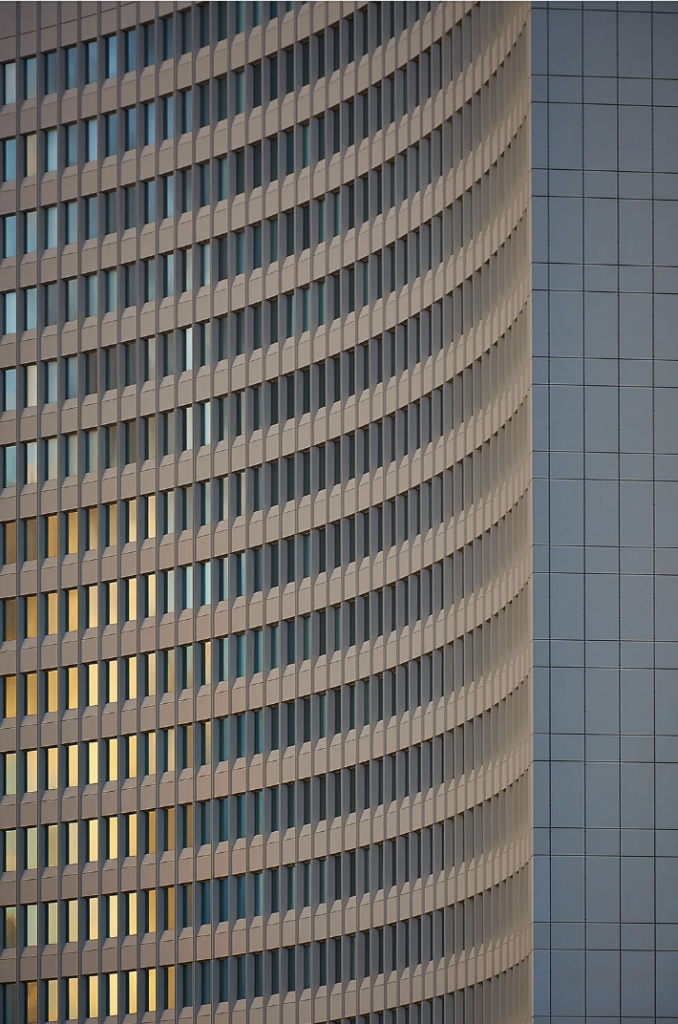 A close-up view of a modern building facade featuring a curved, repeating pattern of windows with vertical lines. The left side of the building's windows are illuminated by golden sunlight, while the right side remains in shadow. Adjacent to the curved structure, a flat wall is visible.