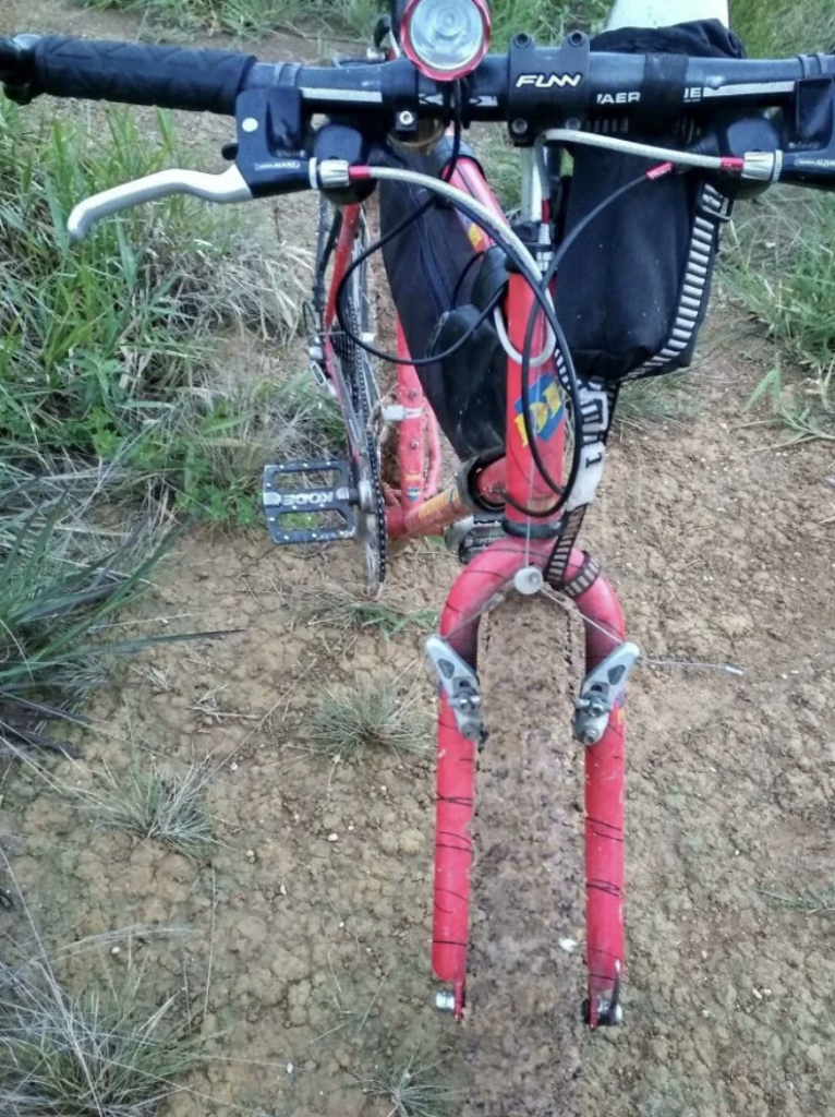 A red bicycle with the front wheel missing is positioned on a dirt path surrounded by grass. The handlebars and pedals are visible, and a black bag is attached to the front frame. The bike appears to be partially disassembled or in need of repair.