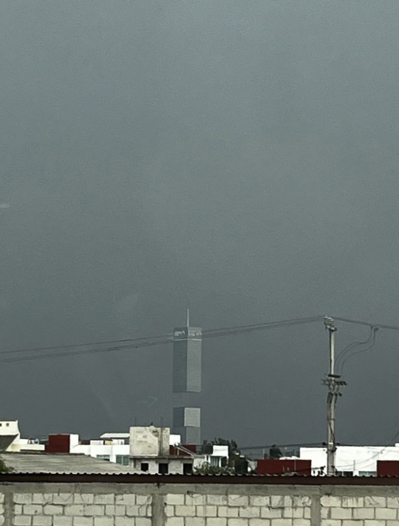 A view of a tall building standing against a dark, stormy sky. The building has a narrow, vertical structure with several stories and a distinct top. The foreground includes lower residential structures, electrical poles, and wires. The overall atmosphere appears gloomy.