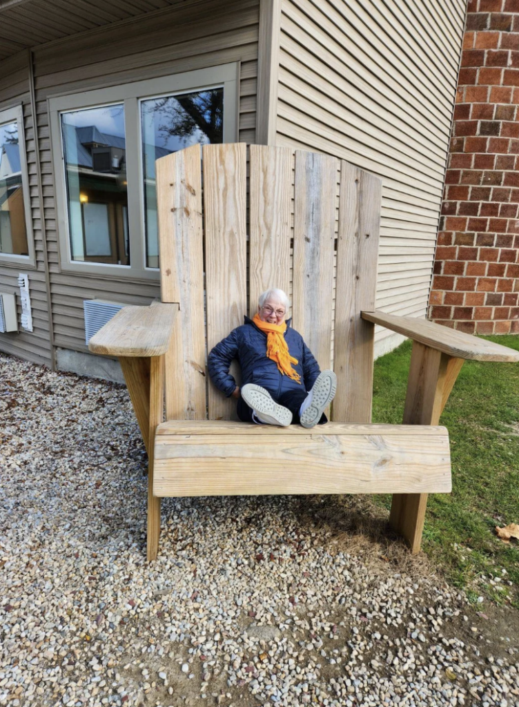 An older woman with white hair and glasses, wearing a blue jacket, yellow scarf, gray pants, and white sneakers, sits in an oversized Adirondack chair in front of a building with beige siding and brick. The chair dwarfs her, emphasizing its large size.