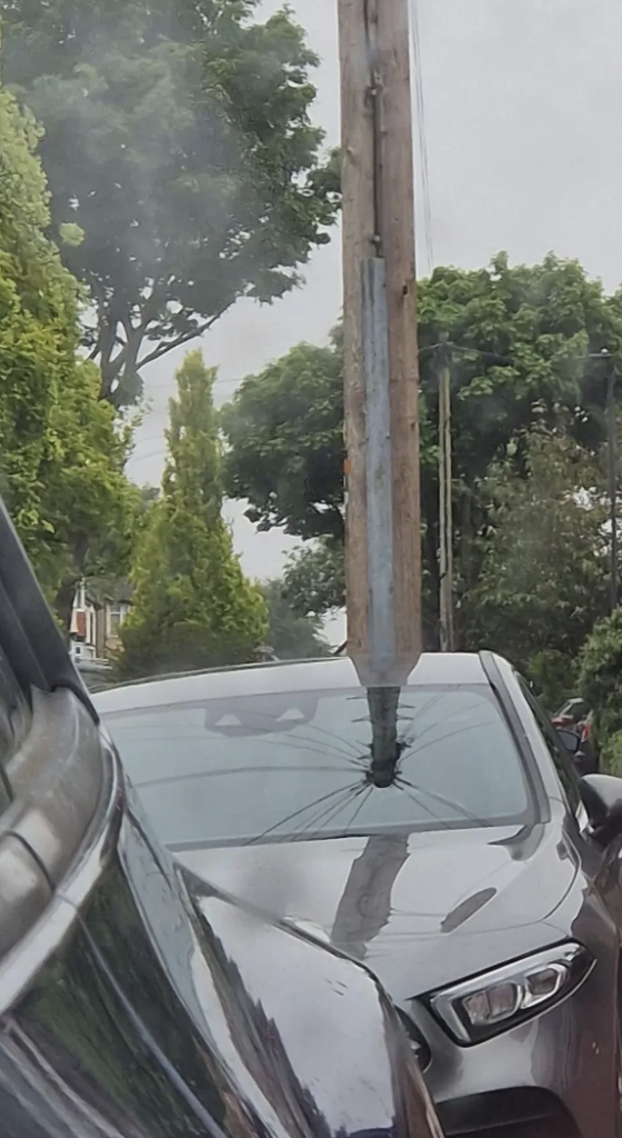 A car with a shattered windshield is parked on a street. A metal pole is lodged into the center of the windshield. Trees and other cars are visible in the background on an overcast day.