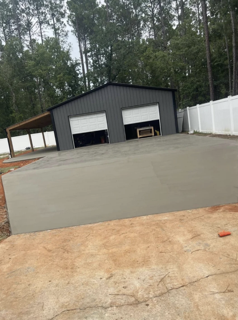 A newly poured concrete driveway leads up to a gray metal garage with two open white doors. The garage has a covered carport section on its left side and is surrounded by a white fence. Tall trees are in the background.