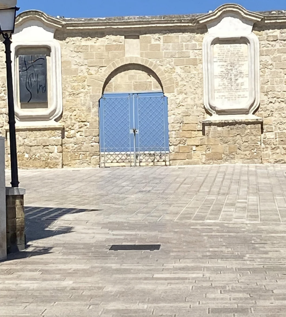 A stone wall with two white plaques and a blue double door at its center. The area in front is a paved courtyard. A lamp post stands on the left edge of the image, casting a shadow on the ground. The sky is clear and blue.