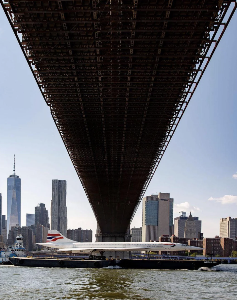 A view from underneath a large bridge overlooking a river, with a ship featuring a British Airways design docked below. The skyline of a city with numerous skyscrapers is visible in the background under a clear blue sky.